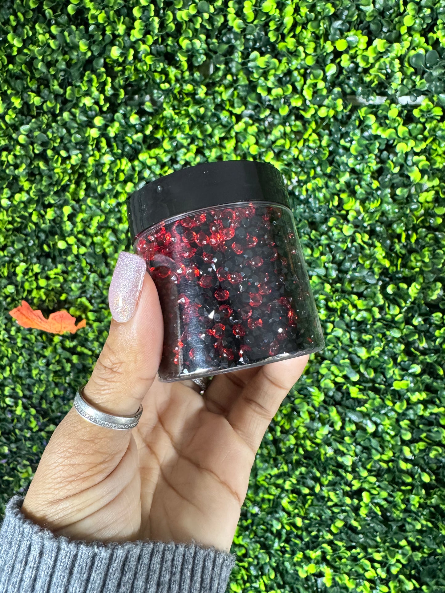 Hand holding a small container of red acrylic crystals against a green grass background