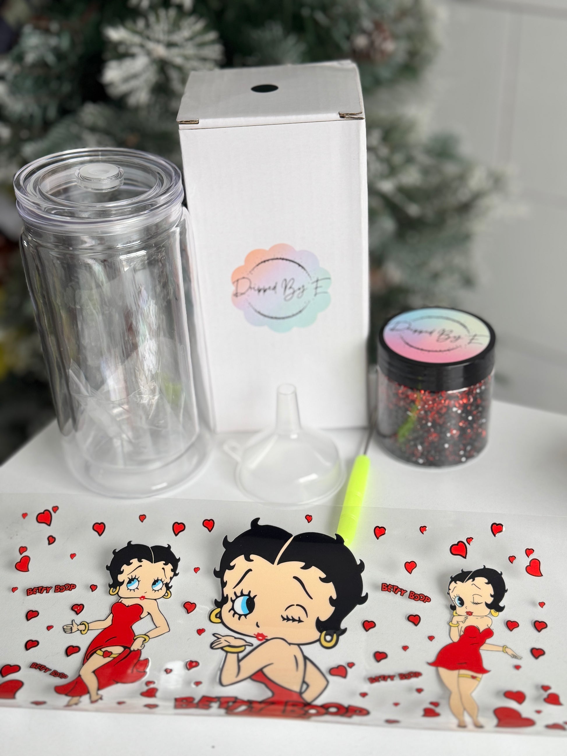 Clear tumbler, white box, funnel, and Betty Boop-themed stickers on a decorative surface with a Christmas tree in the background.